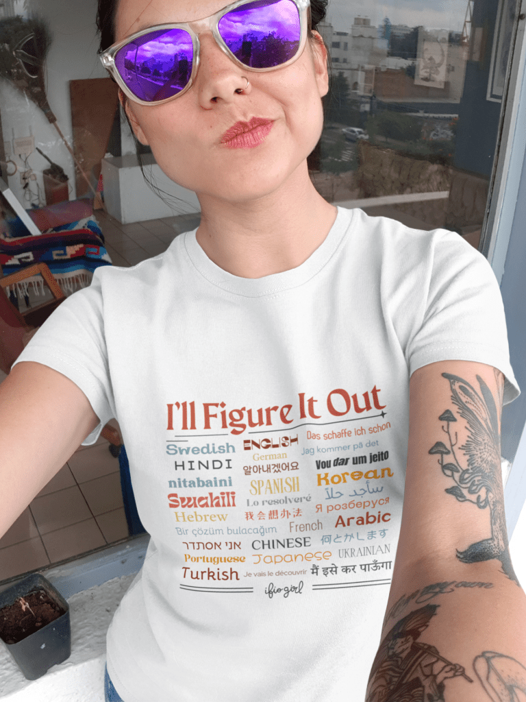 woman wear sunglasses taking a selfie wearing and I'll figure It Out t shirt that has the saying I'll Figure It Out in many different languages around from around the world.