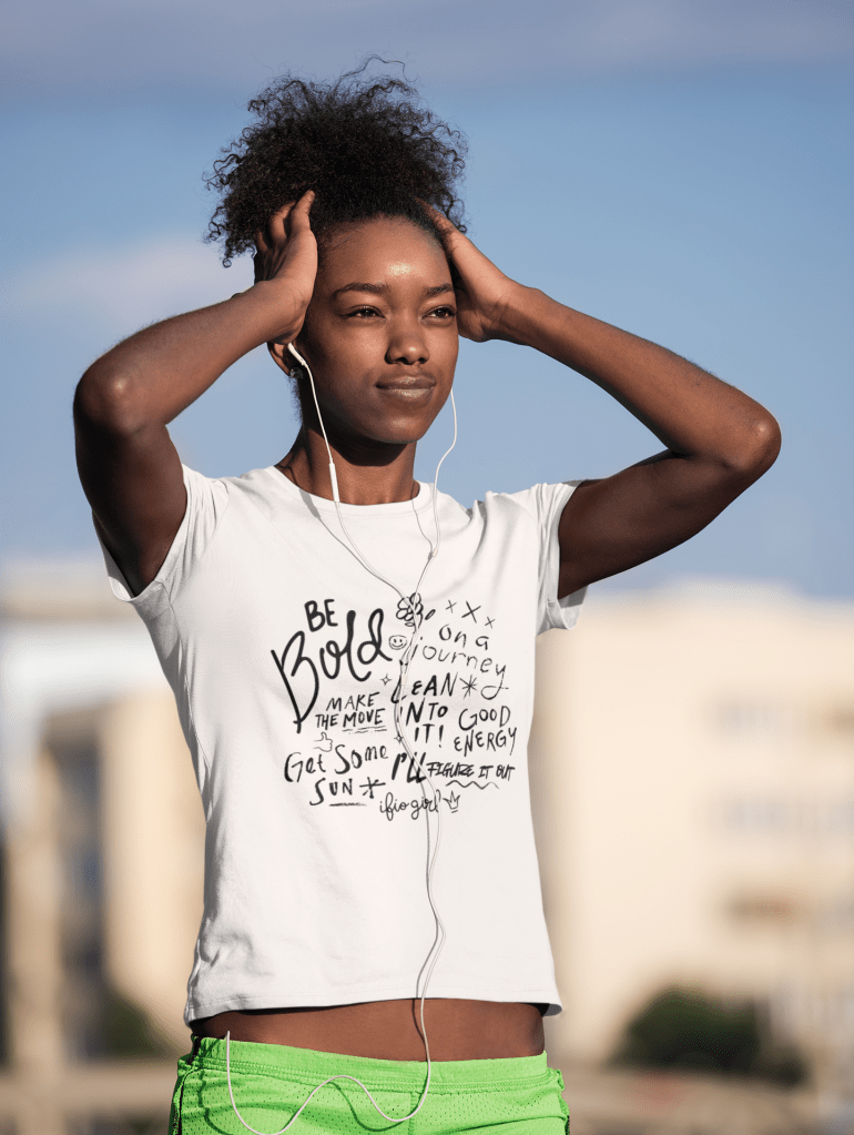 Woman who has ear phones in her ears appears to be working out and wearing a Be Bold doodle t shirt with empowering statements handwritten on the shirt.